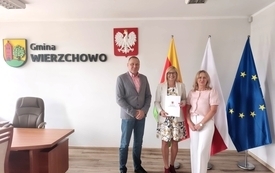 Trzy osoby &ndash; mężczyzna w szarej marynarce oraz dwie kobiety (jedna w jasnym żakiecie i okularach, druga w r&oacute;żowej bluzce) &ndash; siedzą przy długim, drewnianym stole konferencyjnym, na kt&oacute;rym leżą dokumenty i mały bukiet kwiat&oacute;w. W tle, na białej ścianie, widn