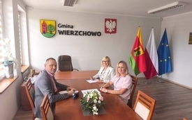 Trzy osoby &ndash; mężczyzna w szarej marynarce oraz dwie kobiety (jedna w jasnym żakiecie i okularach, druga w r&oacute;żowej bluzce) &ndash; siedzą przy długim, drewnianym stole konferencyjnym, na kt&oacute;rym leżą dokumenty i mały bukiet kwiat&oacute;w. W tle, na białej ścianie, widn