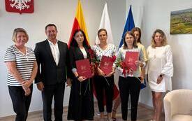 Oficjalne spotkanie w urzędzie Gminy w Wierzchowie. Grupa siedmiu os&oacute;b &ndash; sześć kobiet i jeden mężczyzna &ndash; stoi w rzędzie na tle flag oraz polskiego godła. Trzy kobiety w centrum trzymają czerwone teczki z godłem oraz czerwone r&oacute;że. Wszyscy są elegancko ub