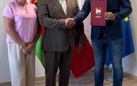 Troje oficjalnie ubranych dorosłych stoi w biurze na tle napisu &bdquo;Gmina WIERZCHOWO&rdquo;, polskiego godła i flag państwowych, a stojący w środku mężczyzna trzyma czerwony dokument.