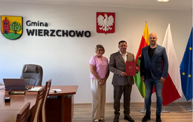 Troje oficjalnie ubranych dorosłych stoi w biurze na tle napisu &bdquo;Gmina WIERZCHOWO&rdquo;, polskiego godła i flag państwowych, a stojący w środku mężczyzna trzyma czerwony dokument.