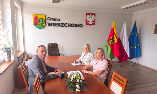 Trzy osoby – mężczyzna w szarej marynarce oraz dwie kobiety (jedna w jasnym żakiecie i okularach, druga w różowej bluzce) – siedzą przy długim, drewnianym stole konferencyjnym, na którym leżą dokumenty i mały bukiet kwiatów. W tle, na białej ścianie, widn