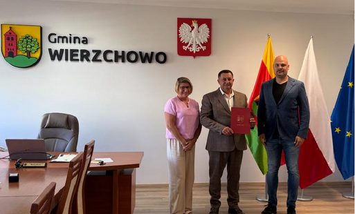 Troje oficjalnie ubranych dorosłych stoi w biurze na tle napisu „Gmina WIERZCHOWO”, polskiego godła i flag państwowych, a stojący w środku mężczyzna trzyma czerwony dokument.